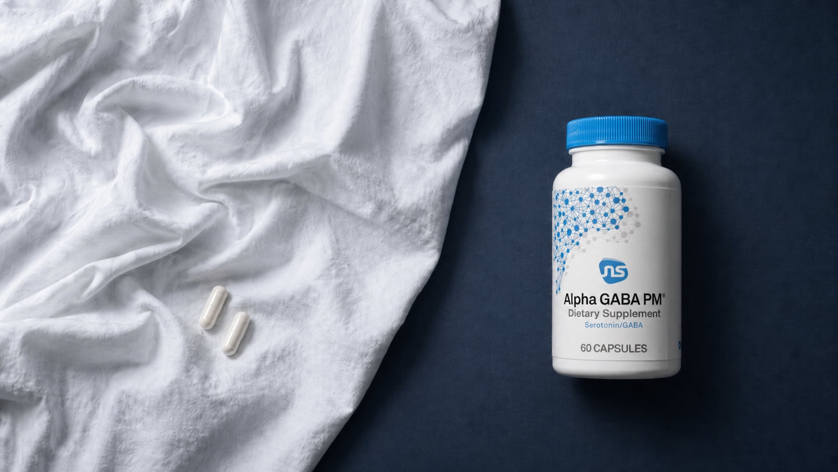 Alpha-Gaba PM bottle on a deep navy surface beside a rumpled white linen bedsheet with two white capsules resting on the fabric