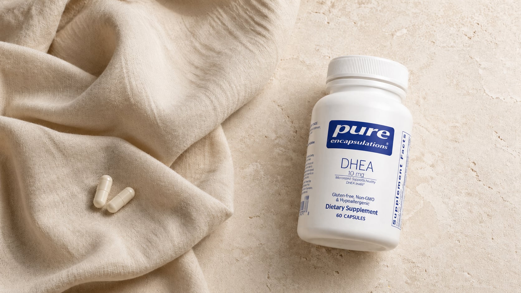 Pure Encapsulations DHEA 10mg bottle on a warm beige stone surface beside a draped cream linen scarf with two white capsules resting on the fabric