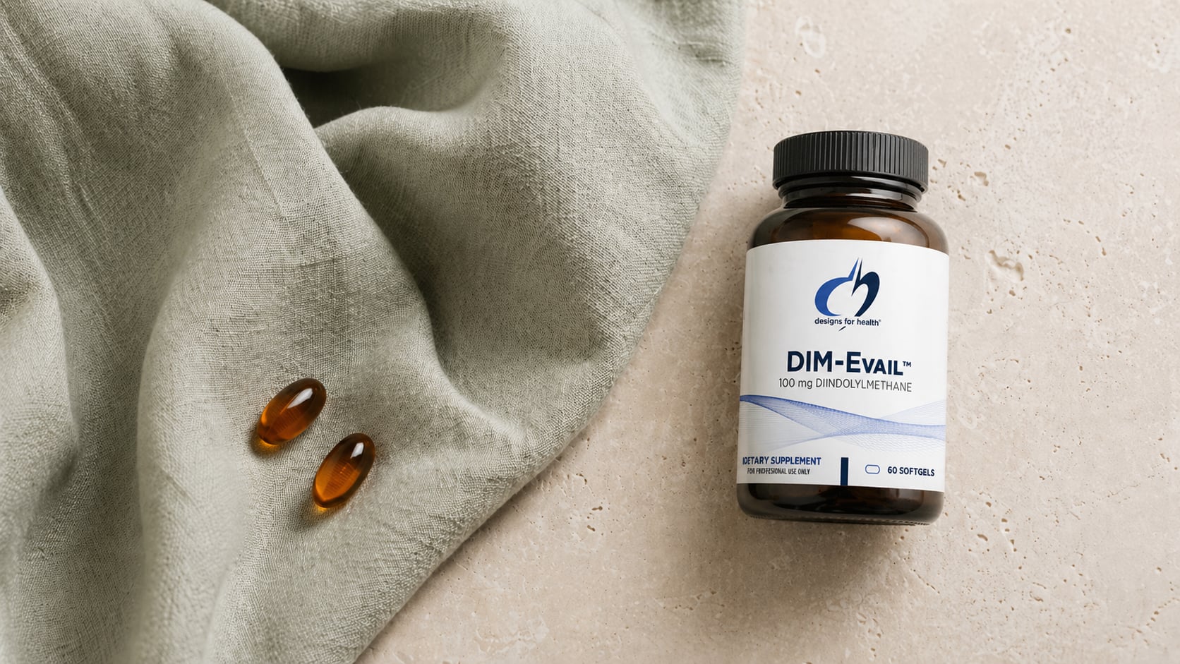 Designs for Health DIM-Evail amber bottle on a warm stone surface beside a draped sage-green linen scarf with two amber softgels resting on the fabric