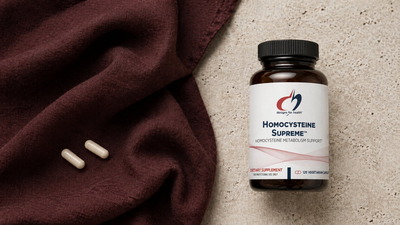 Designs for Health Homocysteine Supreme amber bottle on a warm beige stone surface beside a draped deep-burgundy linen scarf with two white capsules resting on the fabric