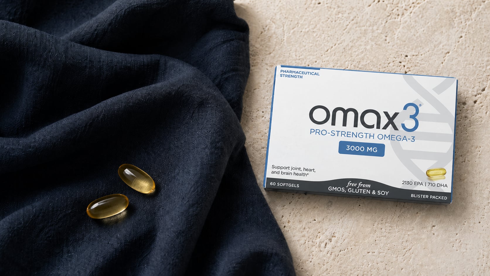 Omax3 Pro-Strength Omega-3 box on a warm beige stone surface beside a draped deep-navy linen scarf with two amber softgels resting on the fabric