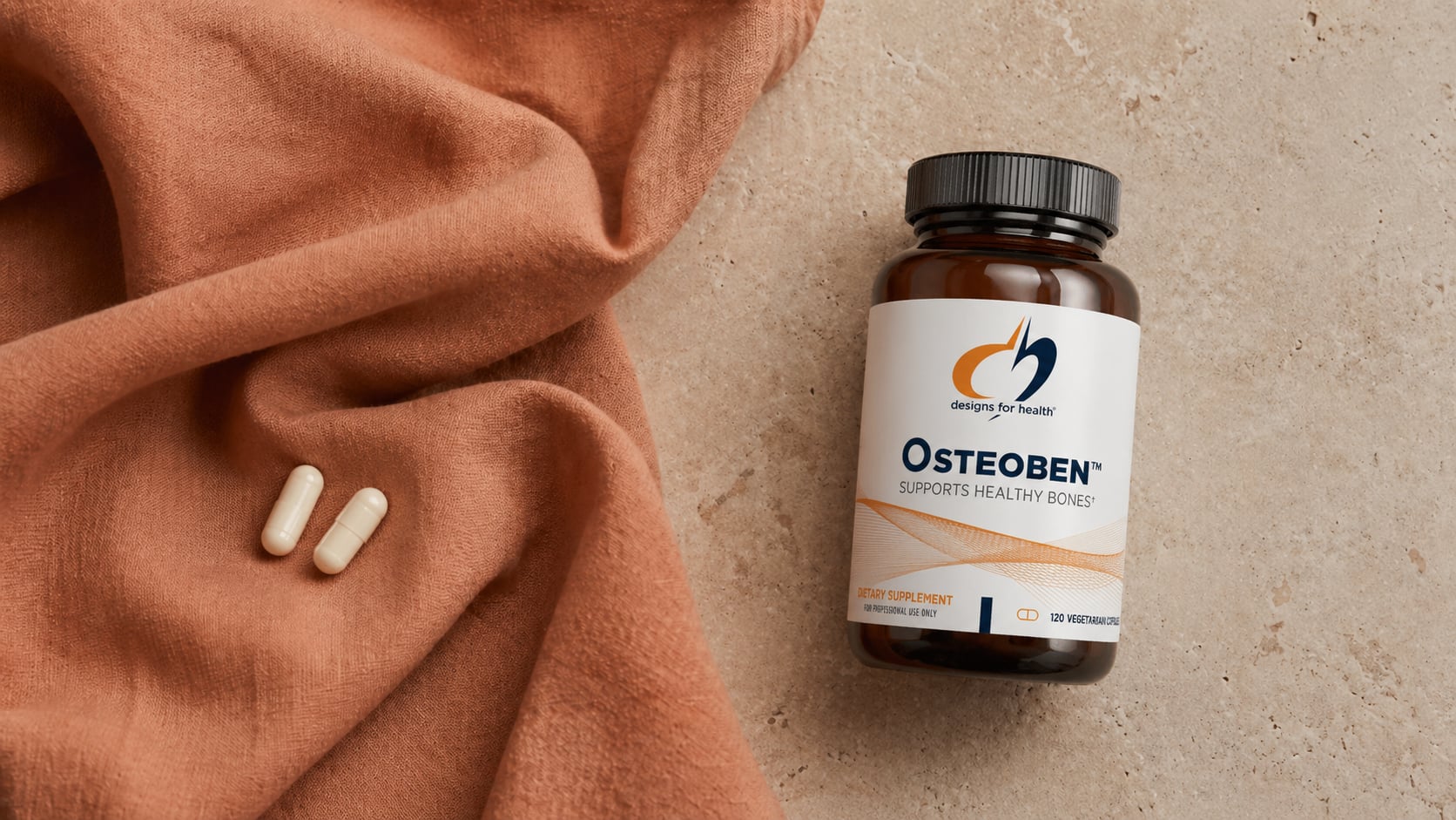 Designs for Health Osteoben amber bottle on a warm beige stone surface beside a draped terracotta linen scarf with two white capsules resting on the fabric