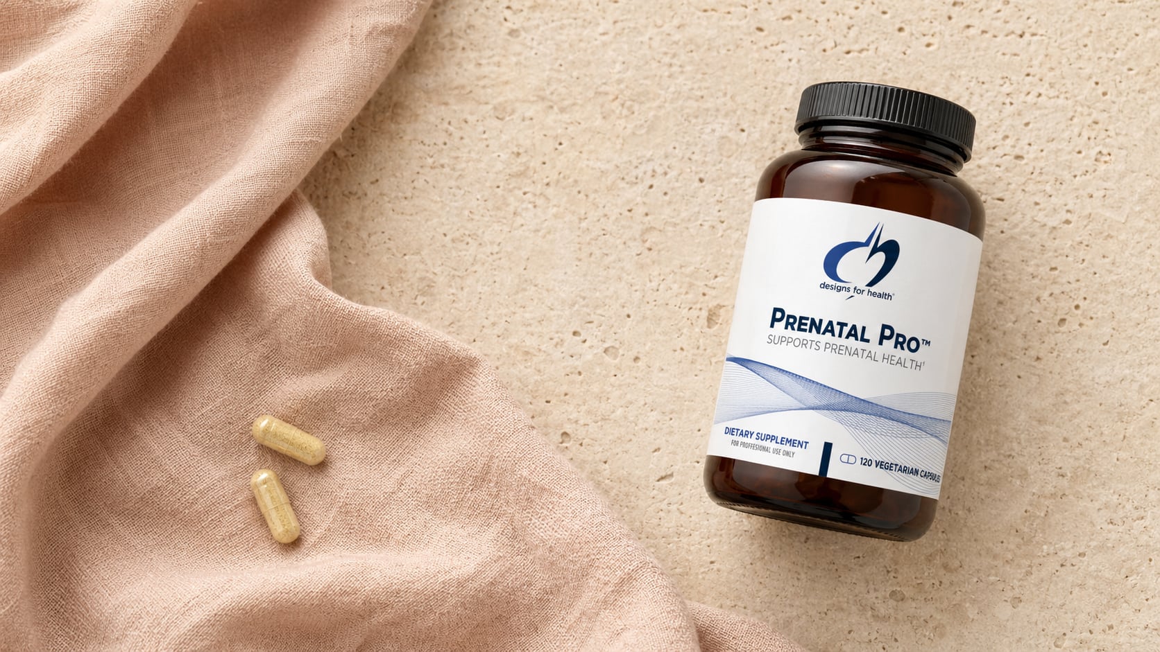 Designs for Health Prenatal Pro amber bottle on a warm beige stone surface beside a draped soft dusty-pink linen scarf with two yellow capsules resting on the fabric