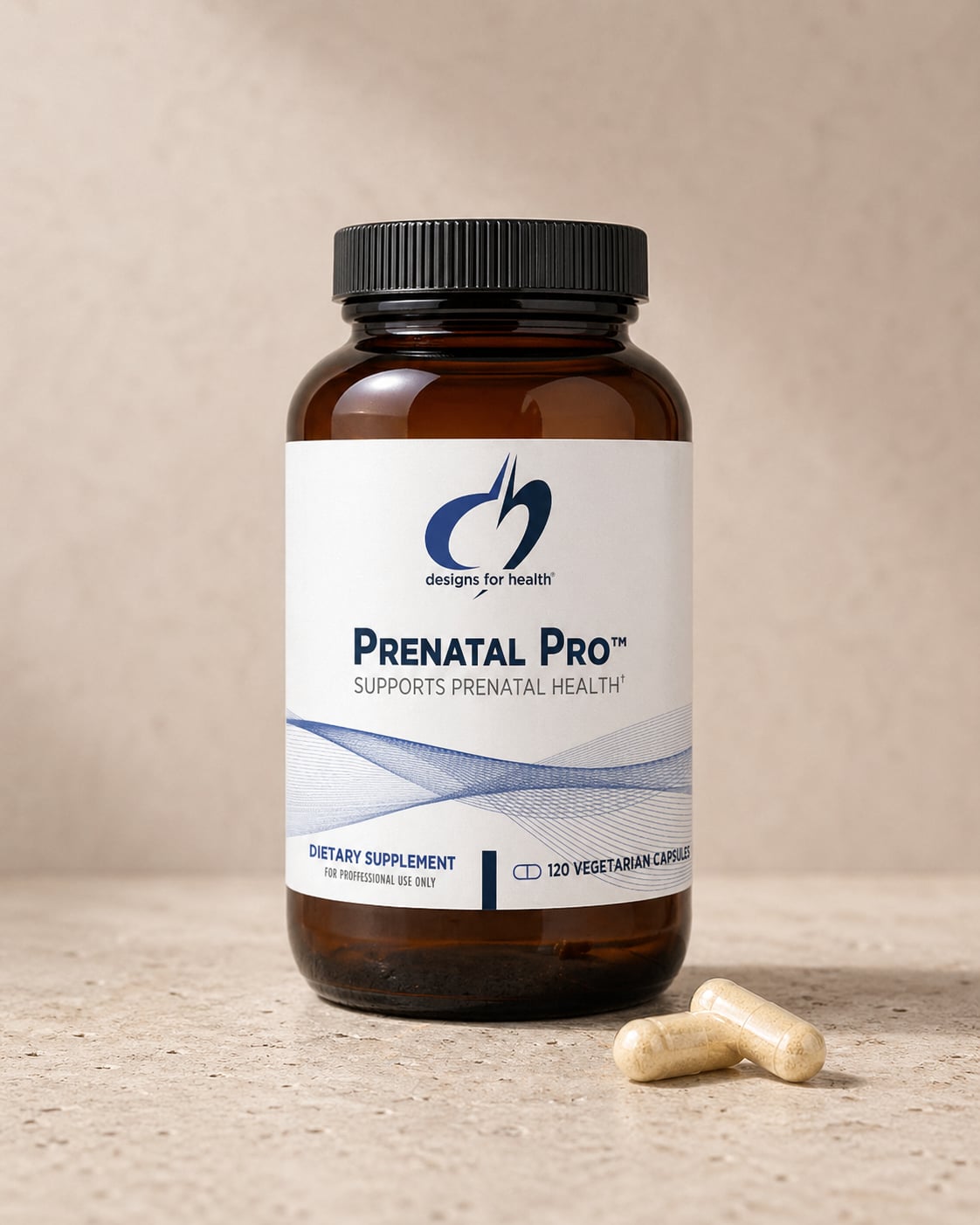 Designs for Health Prenatal Pro amber bottle on a warm beige stone surface against a soft neutral wall, with two white capsules resting at the base