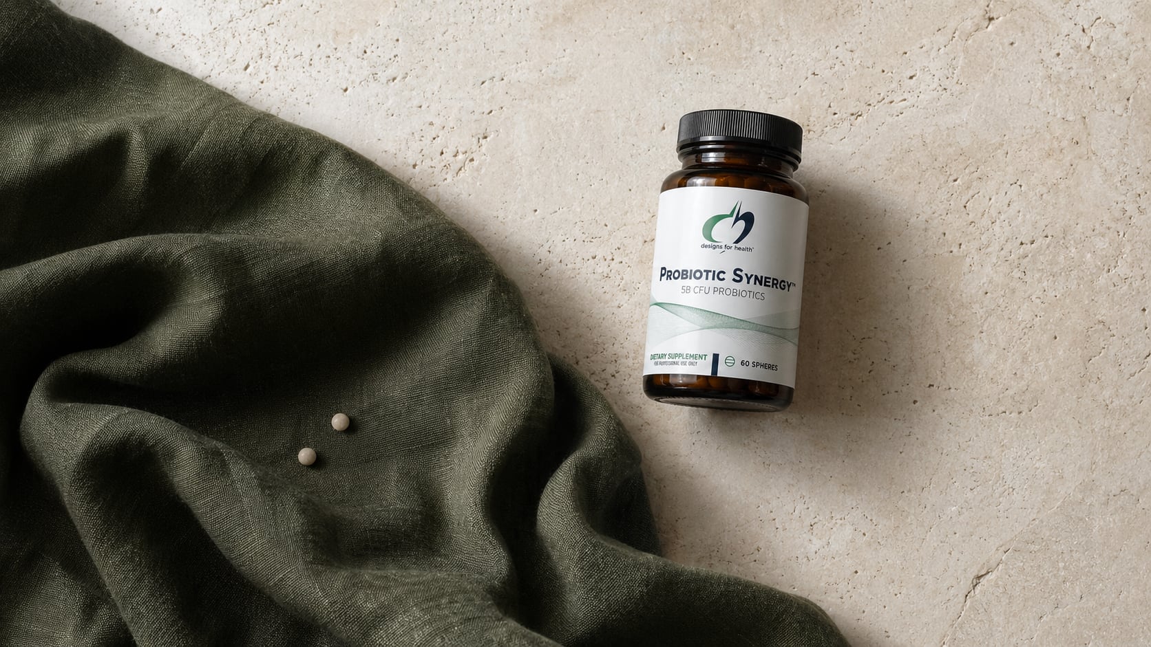 Designs for Health Probiotic Synergy Spheres amber bottle on a warm beige stone surface beside a draped olive-green linen scarf with two small white spheres resting on the fabric