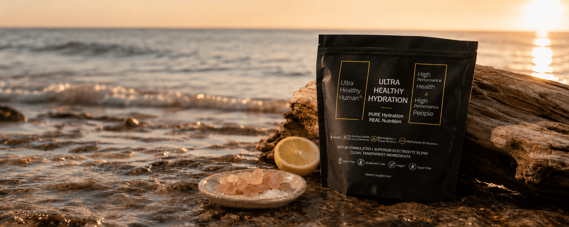 A small ceramic dish of Pink Himalayan salt resting on a sun-warmed rock at the ocean's edge at golden hour, the Ultra Healthy Hydration packet beside it on driftwood