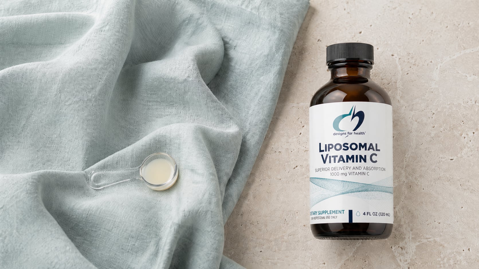 Designs for Health Liposomal Vitamin C amber liquid bottle on a warm beige stone surface beside a draped pale-blue linen scarf with a small dish of the liquid dose