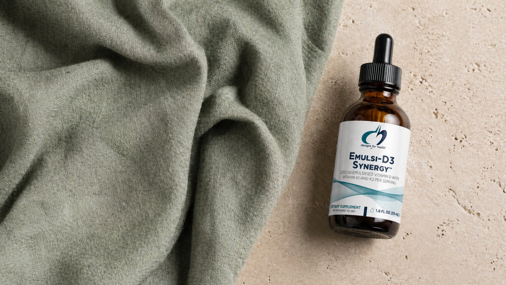 Designs for Health Emulsi-D3 Synergy amber dropper bottle on a warm beige stone surface beside a draped sage-green linen scarf