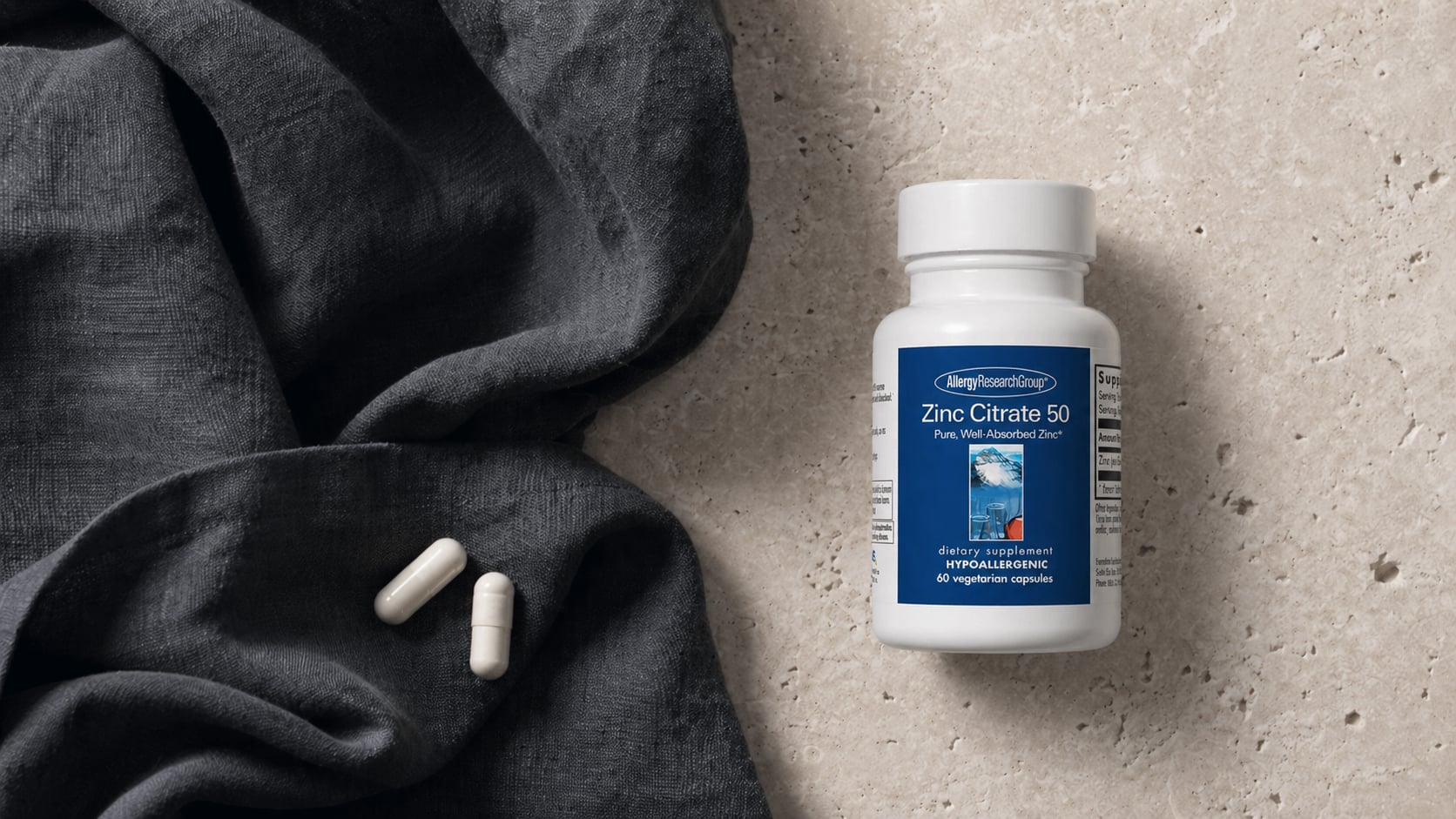 Allergy Research Group Zinc Citrate 50 white bottle on a warm beige stone surface beside a draped charcoal linen scarf with two white capsules resting on the fabric