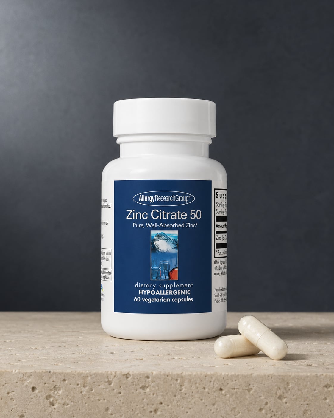 Allergy Research Group Zinc Citrate 50 white bottle on a warm beige stone surface against a deep slate-grey wall, with two white capsules resting at the base